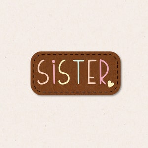 Könnte beinhalten: Ein braunes Leder-Patch mit dem Wort "SISTER" in bunten Buchstaben. In der unteren rechten Ecke befindet sich ein kleines rosa Herz.