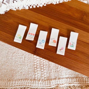 May include: Six small, rectangular fabric tags with floral illustrations and words like "happiness" and "success" printed on them. The tags are arranged on a wooden surface with a cream-colored, textured fabric in the foreground.