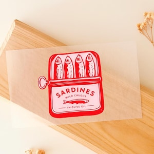 May include: A red and white illustration of a sardine can with four fish inside. The can is labelled "Sardines Wild Caught in Olive Oil". The image is on a light brown paper.