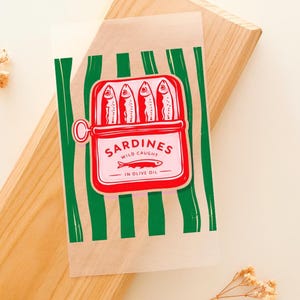 Op de afbeelding: Een doorzichtige cellofaan zak met een rode en witte sardineblik grafiek. Het blik bevat vier vissen en de tekst "Sardines Wild Caught in Olive Oil". De achtergrond heeft groene verticale strepen.