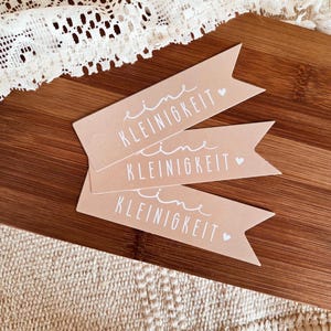 Puede incluir: Tres etiquetas de regalo de color marrón claro con el texto alemán "eine Kleinigkeit" en escritura blanca. Cada etiqueta tiene un pequeño símbolo de corazón. Las etiquetas están dispuestas sobre una superficie de madera.