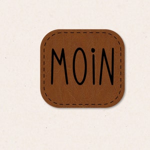 Puede incluir: Un parche de cuero marrón con la palabra "MOIN" escrita en negro.