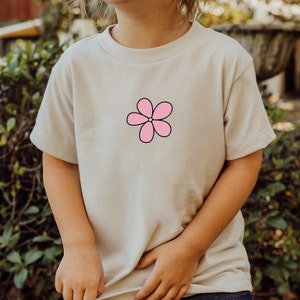 Könnte beinhalten: Ein hellbeiges T-Shirt mit einem pinkfarbenen Blumenmotiv auf der Vorderseite.