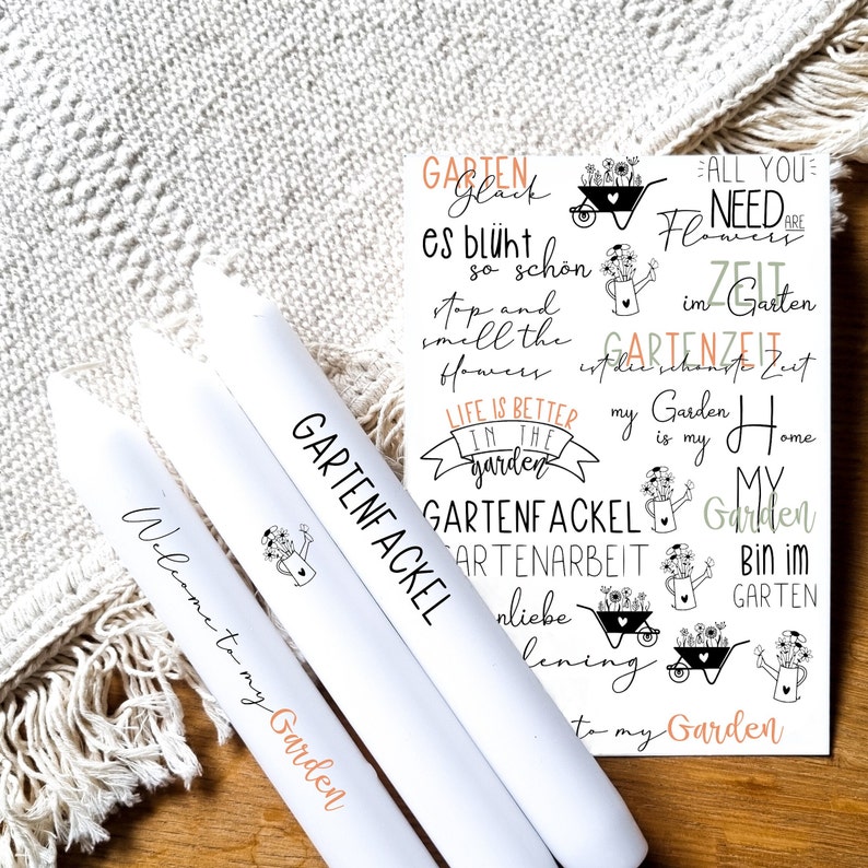 May include: Three white candles with black text. The candles are labeled "Welcome to my Garden", "Gartenfackel", and "Gartenfackel". The background is a white crocheted blanket.