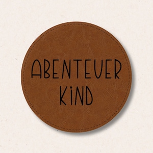 Könnte beinhalten: Ein braunes Leder-Rundetikett mit dem Text "ABENTEUER KIND" in Schwarz.