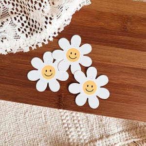 May include: Three white daisy-shaped embellishments with yellow centers and smiling faces. The embellishments are on a wooden surface, with a white lace fabric and a beige woven fabric in the background.