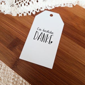 Peut inclure: Une étiquette cadeau blanche avec un haut arrondi et un trou pour l'enfilage. L'étiquette porte les mots "Ein herzliches DANKE" en écriture noire, avec un petit cœur. L'étiquette est sur une surface en bois.