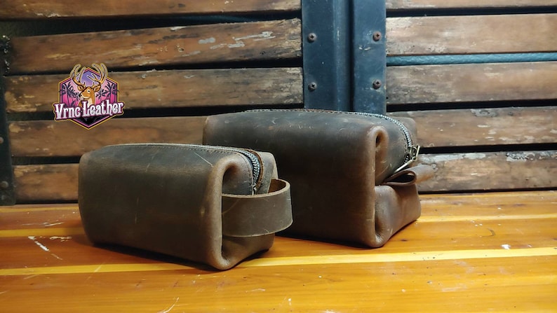 May include: Two brown leather zippered pouches with a brown leather strap on the smaller pouch. The pouches are on a wooden surface.