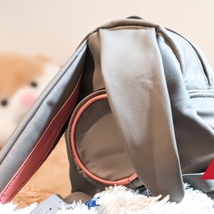 Può includere: Uno zaino in similpelle grigio con una tracolla rosa e un manico rotondo rosa. Lo zaino è appoggiato su una superficie bianca e soffice.
