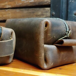 May include: Two brown leather zippered pouches, one larger than the other, are displayed on a wooden surface. The pouches have a distressed look and are made of a thick, durable leather. The larger pouch has a flap that folds over the zipper.