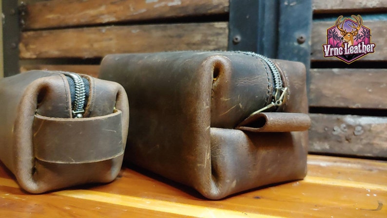 May include: Two brown leather zippered pouches, one larger than the other, are displayed on a wooden surface. The pouches have a distressed look and are made of a thick, durable leather. The larger pouch has a flap that folds over the zipper.