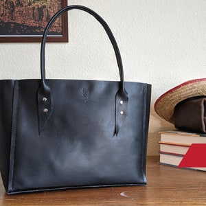 Op de afbeelding: Een zwarte leren tote bag met twee zilveren gespen aan elke kant. De tas staat op een houten tafel. De tekst "PDF" staat in de rechterbenedenhoek van de afbeelding.