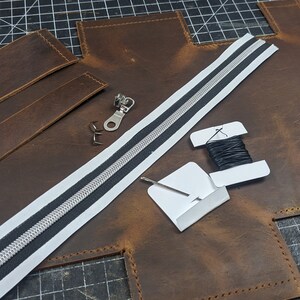 May include: A leather craft project with brown leather pieces, a white zipper with a black stripe, black thread, and a metal zipper pull.