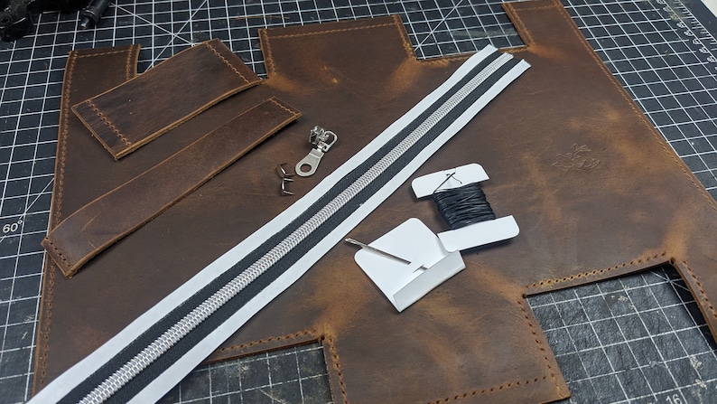 May include: A leather craft project with brown leather pieces, a white zipper with a black stripe, black thread, and a metal zipper pull.