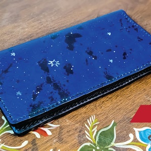 Peut inclure: Un portefeuille en cuir bleu avec un motif de ciel étoilé. Le portefeuille est fermé et posé sur une surface en bois avec des fleurs peintes.