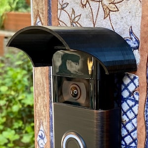 May include: A bronze-colored Ring video doorbell with a black protective cover, mounted on a decorative pillar. The doorbell features a camera lens and a circular button with a white light. The pillar is adorned with patterned tiles.