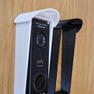 May include: A black Eufy Security video doorbell with a white protective cover is mounted on a wooden surface. The text "Remove the film before use" is visible. A black bracket is also shown.