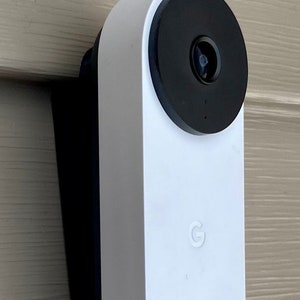 May include: A white Google Nest Doorbell with a black camera lens and a white button. The doorbell is mounted on a light brown wall.