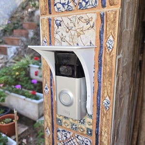 May include: A silver Ring video doorbell mounted on a decorative tiled pillar. The doorbell is installed with a white protective cover. The pillar is decorated with blue, brown, and white tiles, and the word "ring" is visible on the doorbell.