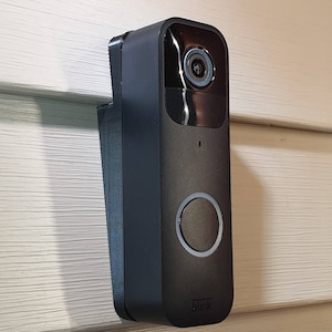 May include: A black doorbell camera with a wide-angle lens and a button for visitors to press. The camera is mounted on a white wall and has the word "blink" printed on the bottom.