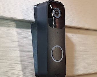 May include: A black doorbell camera with a wide-angle lens and a button for visitors to press. The camera is mounted on a white wall and has the word "blink" printed on the bottom.