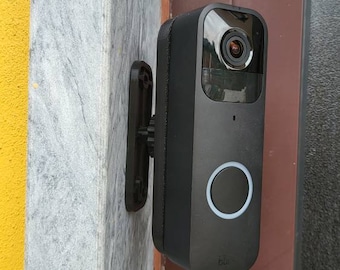 May include: A black smart doorbell with a camera lens and a white illuminated ring around a button. The doorbell is mounted on a wall with a dark brown bracket. The wall is a mix of gray and yellow.