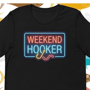 May include: Black t-shirt with the text "WEEKEND HOOKER" in a neon-style design, featuring a fishing hook and worm. The shirt is set against a background of colourful fish illustrations.