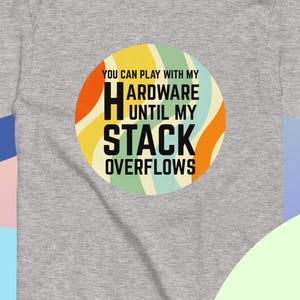 May include: A heather gray t-shirt with a circular graphic. The graphic features the text "YOU CAN PLAY WITH MY HARDWARE UNTIL MY STACK OVERFLOWS" in black and yellow, with orange, green, and blue accents. The shirt is a short-sleeved tee.