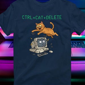 May include: Navy blue t-shirt with the text "CTRL+CAT+DELETE" above a cartoon illustration of an orange cat jumping over a broken computer. The design is printed on a dark blue tee.
