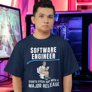 May include: Navy blue t-shirt with the text "SOFTWARE ENGINEER" in white. Below the text is a graphic of a person on a toilet with a tablet. The text "STARTS EVERY DAY WITH A MAJOR RELEASE" is below the graphic.