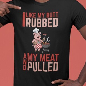 May include: Black t-shirt with the text "LIKE MY BUTT RUBBED AND MY MEAT PULLED" in white and red lettering. The shirt features a cartoon pig chef grilling sausages and holding tongs. A fun design for barbecue enthusiasts.