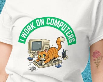 Peut inclure: T-shirt blanc avec un dessin animé d'un chat tigré orange sur un clavier, un ordinateur rétro et le texte "I WORK ON COMPUTERS" en vert. Le chat interagit avec le clavier. Une souris et d'autres accessoires informatiques sont également visibles.