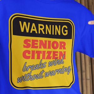 May include: Royal blue t-shirt with a yellow and black warning sign graphic. The sign reads "WARNING SENIOR CITIZEN breaks wind without warning" in bold, colorful text. The shirt is a casual style.