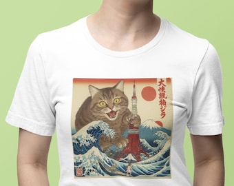 Puede incluir: Camiseta blanca con un diseño de inspiración japonesa que representa un gato gigante atacando la Torre de Tokio. El diseño incluye un sol rojo, olas azules y el texto "大怪獣ジラ". El gato es marrón y gris con ojos amarillos.