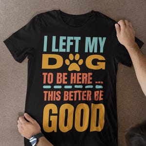 Puede incluir: Camiseta negra con el texto "I LEFT MY DOG TO BE HERE... THIS BETTER BE GOOD" en verde azulado, naranja y dorado. La palabra "DOG" presenta una huella de pata. Un diseño casual y humorístico para amantes de los perros.
