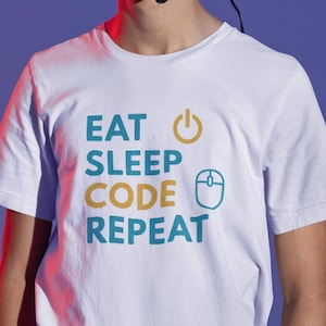 Può includere: T-shirt bianca con la scritta "EAT SLEEP CODE REPEAT" in verde acqua e oro. Il design include un pulsante di accensione e un'icona del mouse del computer. La maglietta è un capo casual.