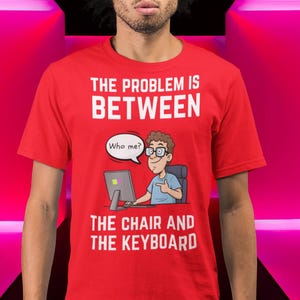 Peut inclure: T-shirt rouge avec texte blanc : « LE PROBLÈME EST ENTRE LA CHAISE ET LE CLAVIER ». Un dessin animé montre une personne devant un ordinateur avec une bulle de dialogue disant « Qui moi ? » La personne porte des lunettes et pointe.