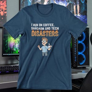 Könnte beinhalten: Marineblaues T-Shirt mit dem Text "I RUN ON COFFEE, SARCASM AND TECH DISASTERS". Das Shirt zeigt eine Cartoon-Grafik einer Person, die eine Kaffeetasse und einen Bleistift hält. Das Design ist in Weiß und Orange gehalten.