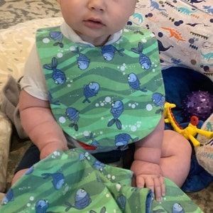 May include: A baby wearing a green and blue bib with a fish pattern. The bib has a white border and is made of soft fabric. The baby is sitting on a colourful rug with a dinosaur pattern.