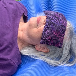 May include: A person wearing a purple shirt lies on a blue yoga mat with a purple and pink floral eye mask covering their eyes.