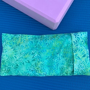 May include: A rectangular eye pillow with a teal and green leaf pattern, next to a lavender yoga block, on a blue surface. The eye pillow is made of fabric and has a folded edge.