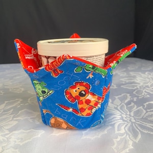 May include: A blue fabric ice cream bowl cosy with a red interior. The cosy features a playful dog print with the words "Woof" and "Bark". The cosy is designed to hold a pint-sized ice cream container.