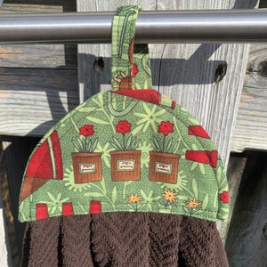 May include: A brown kitchen towel with a decorative top featuring a green floral pattern and three flower pots labelled "Green Gardens." The top has a red border and a loop for hanging.
