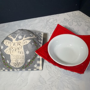 May include: A white ceramic bowl rests on a red quilted bowl cosy. A grey bowl cosy with a deer design and the text "Over the River and Through the Woods" is also present. The bowl cosies are on a white floral patterned surface.