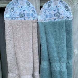 May include: Two hanging kitchen towels with a light blue fabric top featuring a pattern of sea turtles, shells, and coral. One towel is a solid beige colour, and the other is a solid teal colour. Both towels have a loop for hanging on a rod.