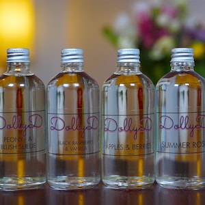 May include: Four clear glass bottles with silver lids and black labels. The labels have the text "Dolly D" in pink script and the scent names: "Peony & Blush Suede", "Black Raspberry & Vanilla", "Apples & Berries", and "Summer Rose".