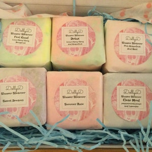 K&ouml;nnte beinhalten: Eine Schachtel mit sechs Dusch-Dampfern in verschiedenen Duftrichtungen. Die Dusch-Dampfer sind in wei&szlig;es Papier mit rosa und wei&szlig;en Etiketten verpackt. Die Etiketten tragen den Text "Dolly D Shower Steamer" und den Duft jedes Dusch-Dampfers. Die D&uuml;fte sind Feel Good, Detox, Pink Grapefruit und Basilikum, Sweet Dreams, Sommerrose und Clear Mind.