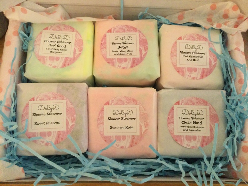 K&ouml;nnte beinhalten: Eine Schachtel mit sechs Dusch-Dampfern in verschiedenen Duftrichtungen. Die Dusch-Dampfer sind in wei&szlig;es Papier mit rosa und wei&szlig;en Etiketten verpackt. Die Etiketten tragen den Text "Dolly D Shower Steamer" und den Duft jedes Dusch-Dampfers. Die D&uuml;fte sind Feel Good, Detox, Pink Grapefruit und Basilikum, Sweet Dreams, Sommerrose und Clear Mind.
