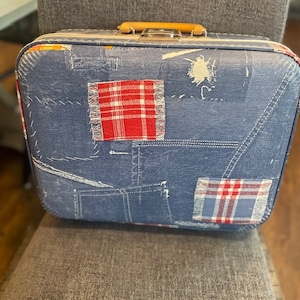 Pode incluir: Uma mala vintage com um padrão semelhante ao denim e remendos xadrez vermelhos. A mala tem uma alça de madeira e fechos de metal. O design geral é em estilo patchwork, com detalhes de costura visíveis.