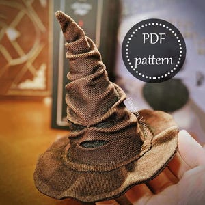 Puede incluir: Un sombrero seleccionador marrón y texturizado con una parte superior puntiaguda y un ala ancha. El sombrero tiene un aspecto desgastado y está hecho de una tela acanalada. Un círculo negro con las palabras "PDF pattern" está en el fondo.